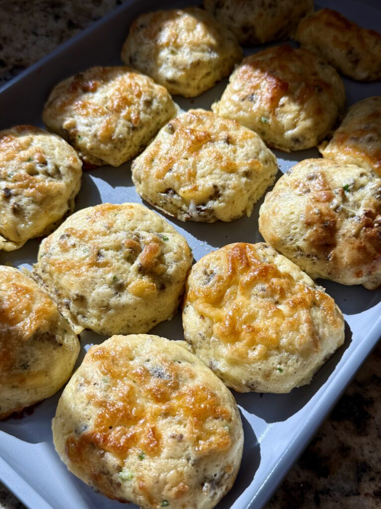 A tray of golden-brown, homemade savory biscuits with herbs and cheese, fresh from the oven, sits on a countertop with sunlight highlighting their texture.