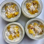 Four glass jars filled with chia pudding, topped with diced mango and shredded coconut, arranged on a white surface.