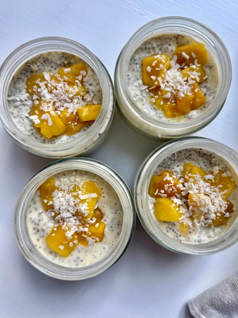 Four glass jars filled with chia pudding, topped with diced mango and shredded coconut, arranged on a white surface.