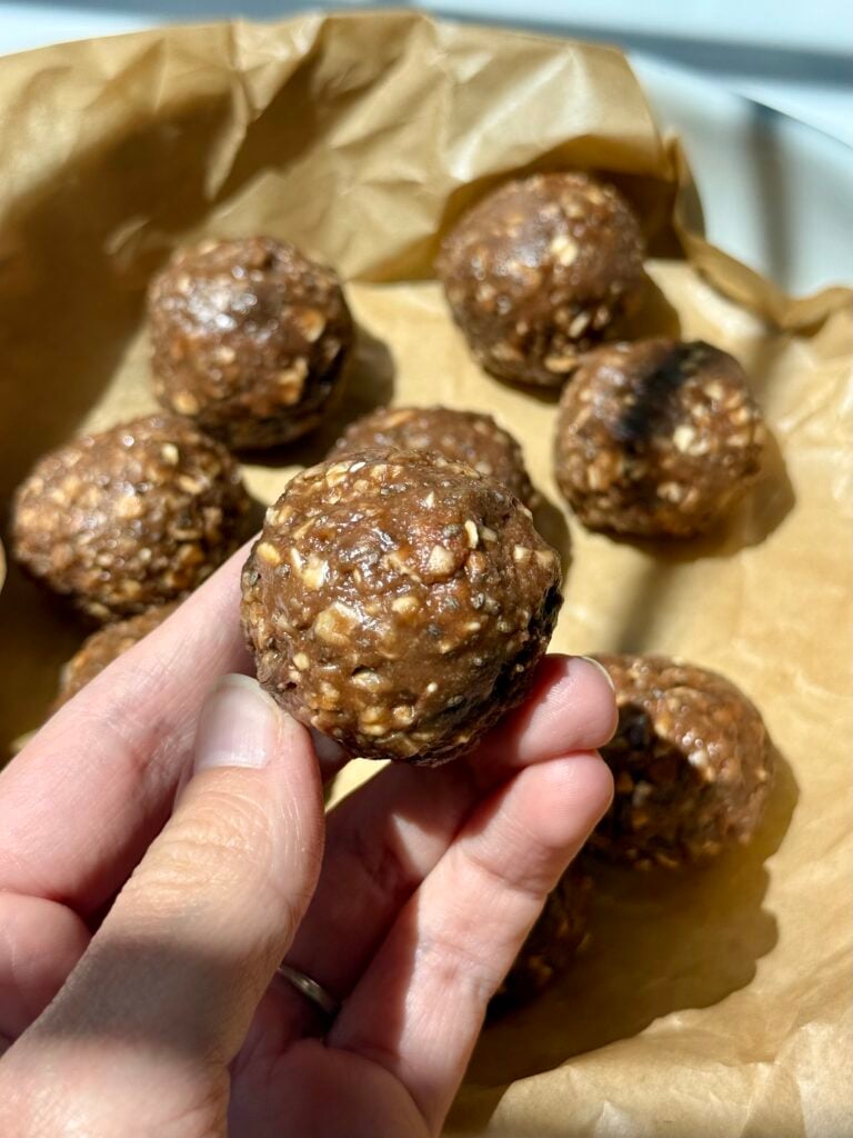 A hand holds a round chocolate energy bite with oats above several more bites resting on crumpled brown parchment paper in a dish.