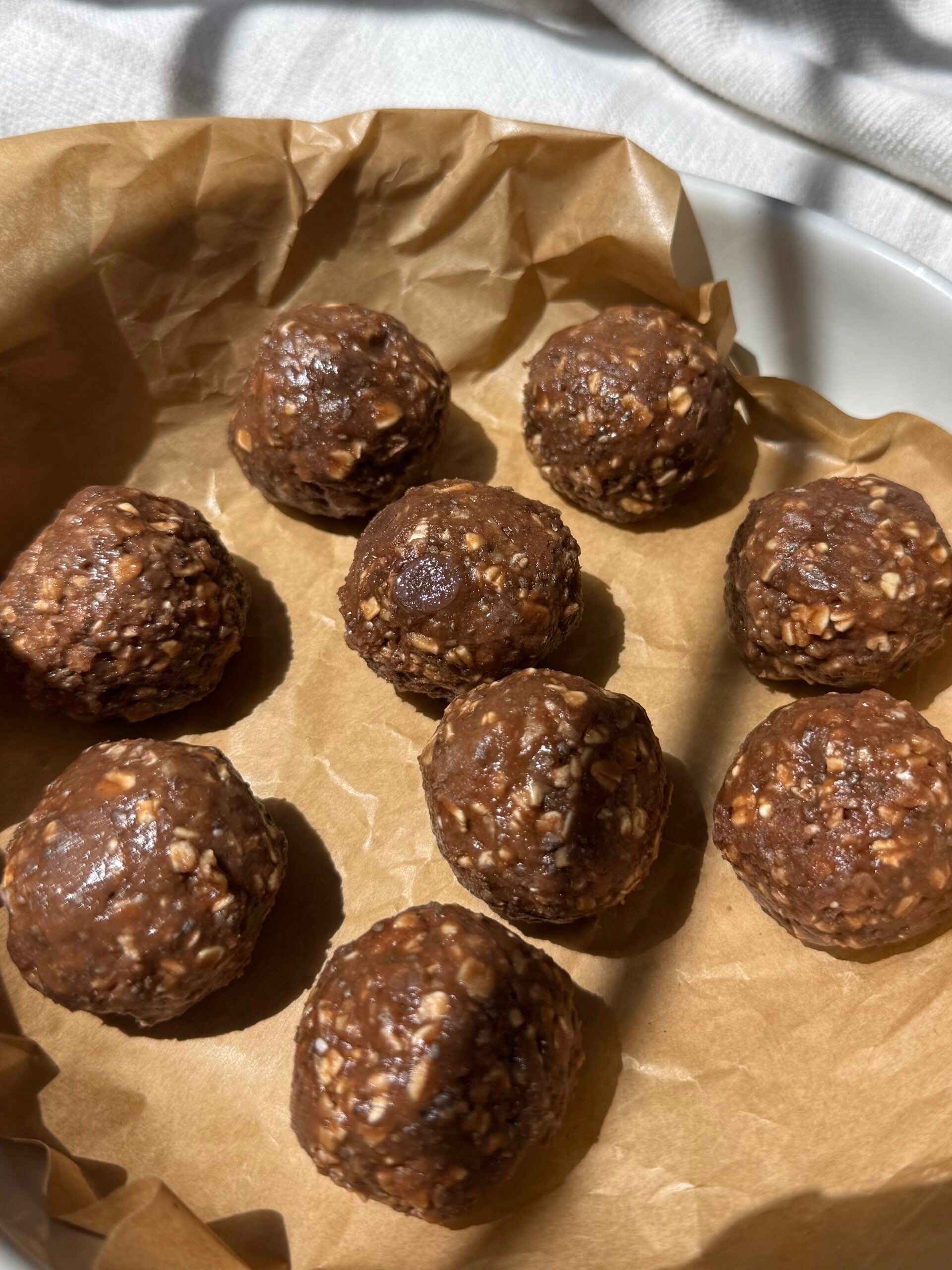 Nine round chocolate oat energy bites on brown parchment paper in a white dish, with sunlight casting soft shadows.