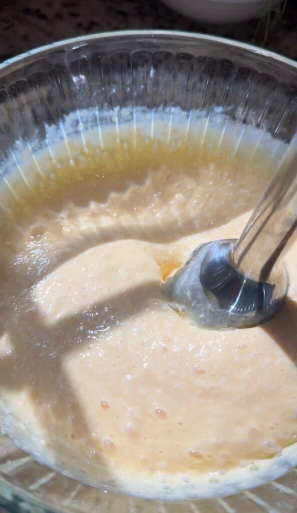 A high-angle close-up shows a stainless steel immersion blender mixing a pale orange, thick liquid batter inside a glass bowl, with visible bubbles and texture throughout the creamy mixture.