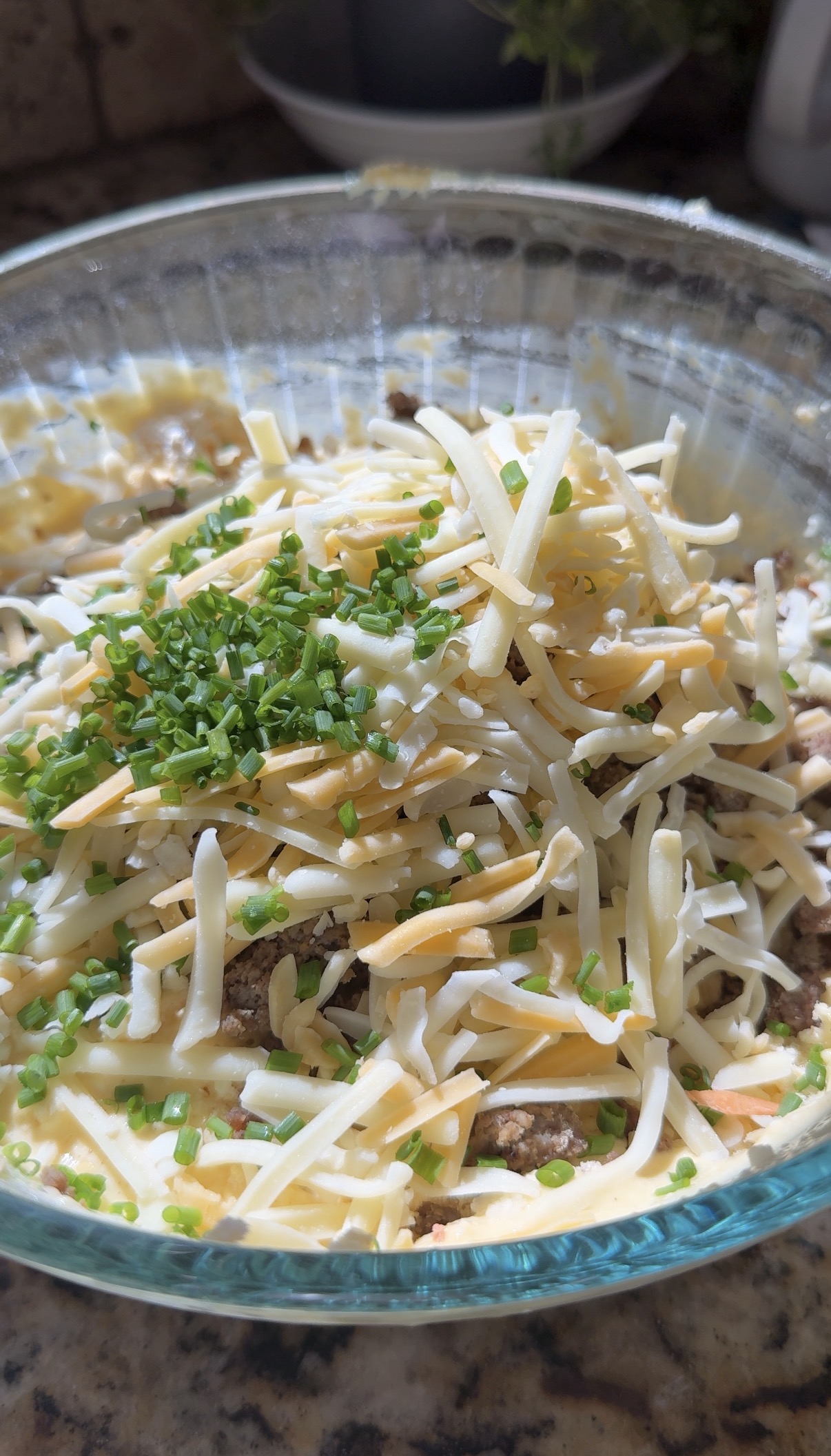A close-up of a glass bowl filled with shredded cheese, chopped chives, ground meat, and creamy sauce, ready to be mixed or used as a casserole filling.
