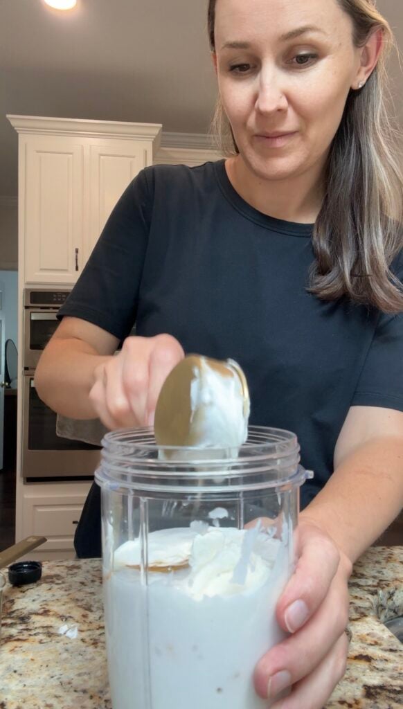 A woman in a black shirt scoops white yogurt from a spoon into a blender cup in a kitchen with white cabinets.