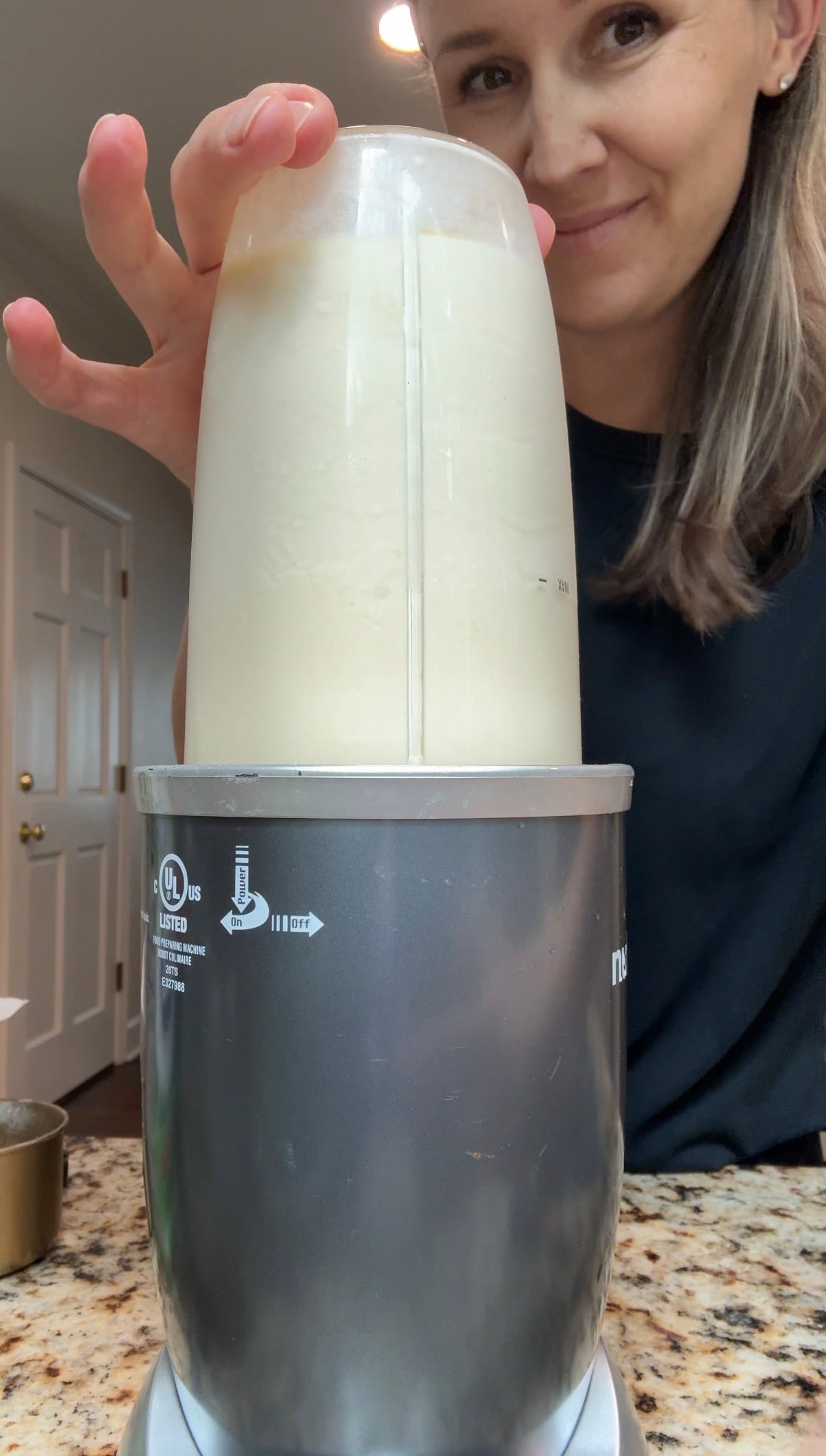 A woman blending a light-colored smoothie in a NutriBullet blender on a kitchen counter, with her hand resting on top of the blender cup.