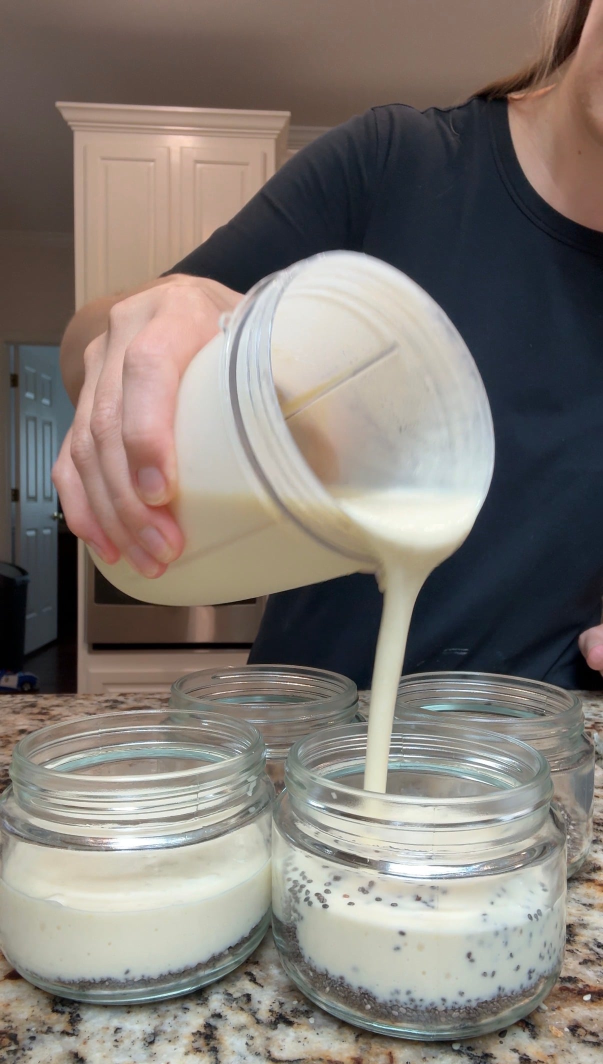 A person pours a creamy liquid from a blender cup into glass jars on a kitchen counter, with three jars visible in the foreground.
