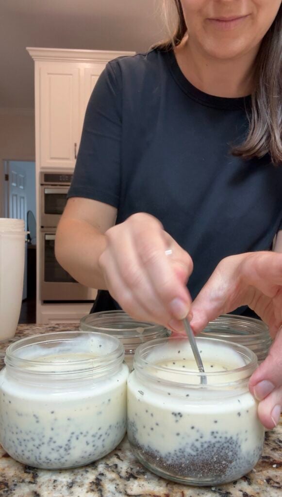 A woman in a black shirt stirs a creamy mixture with black specks in glass jars on a kitchen counter, with more jars and a white container nearby.