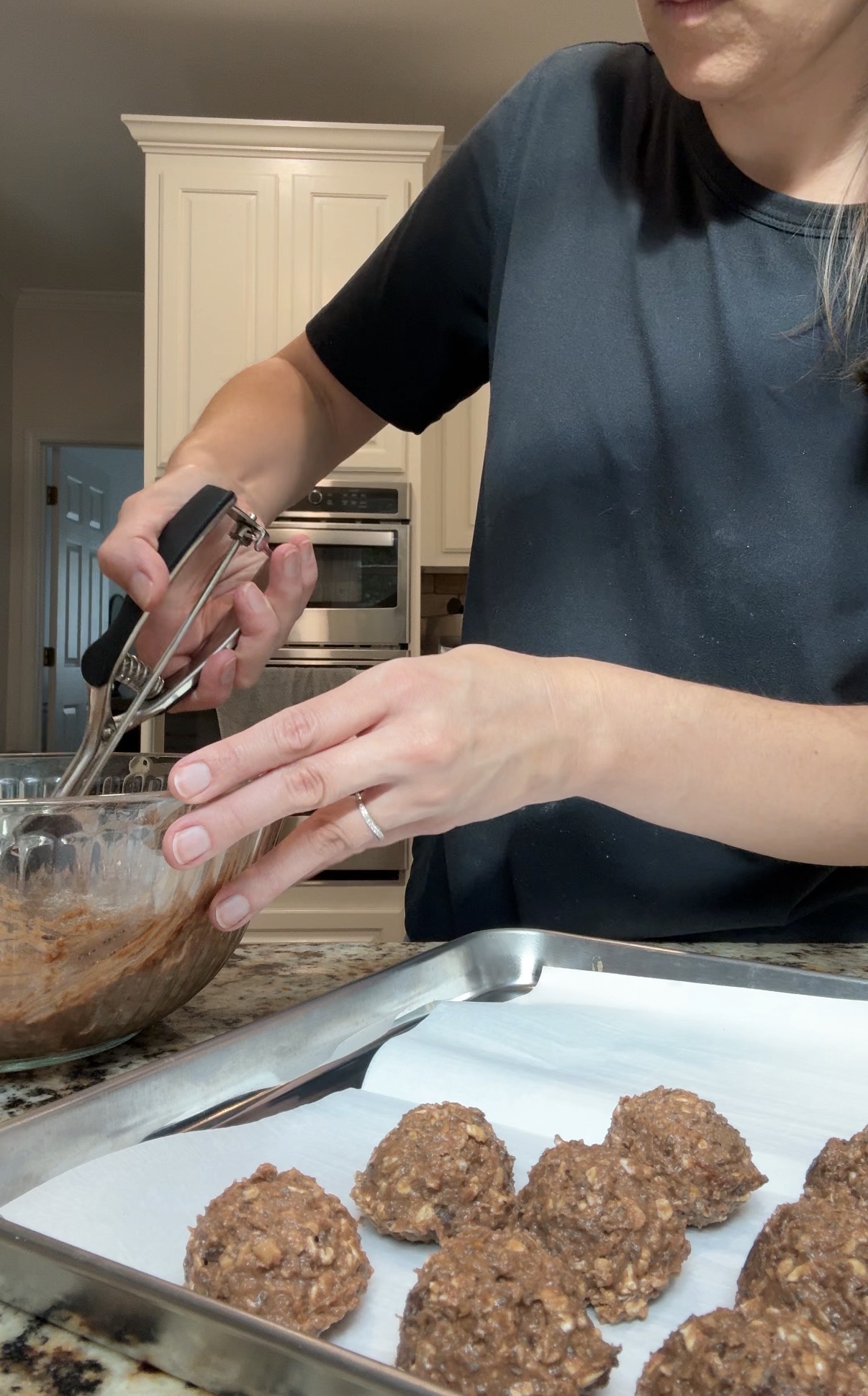 A person uses a cookie scoop to portion dough onto a parchment-lined baking tray in a kitchen.