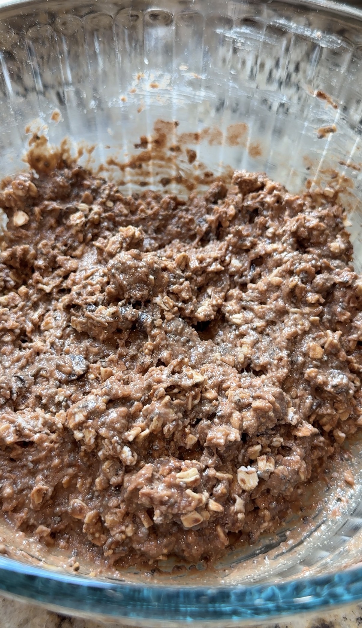 A glass bowl filled with a chunky mixture of oats, cocoa, and chocolate dessert, on a kitchen countertop.
