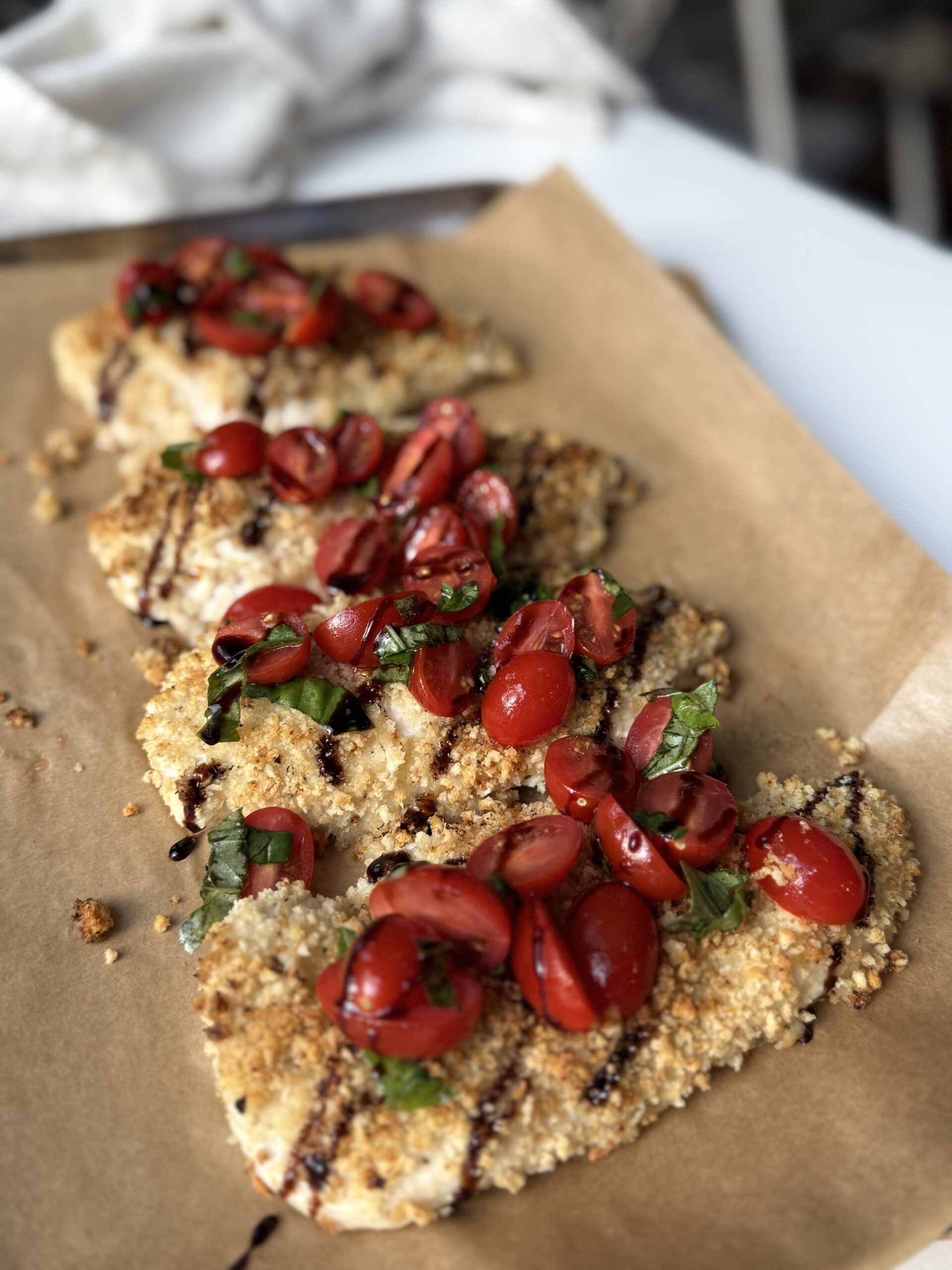 Breaded chicken breasts topped with halved cherry tomatoes, fresh basil, and a drizzle of balsamic glaze, served on parchment paper.