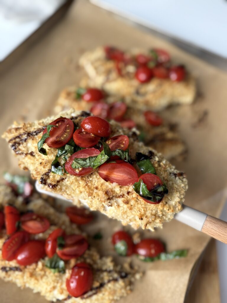 A breaded chicken cutlet topped with sliced cherry tomatoes and fresh basil is held above a baking tray with more cutlets.