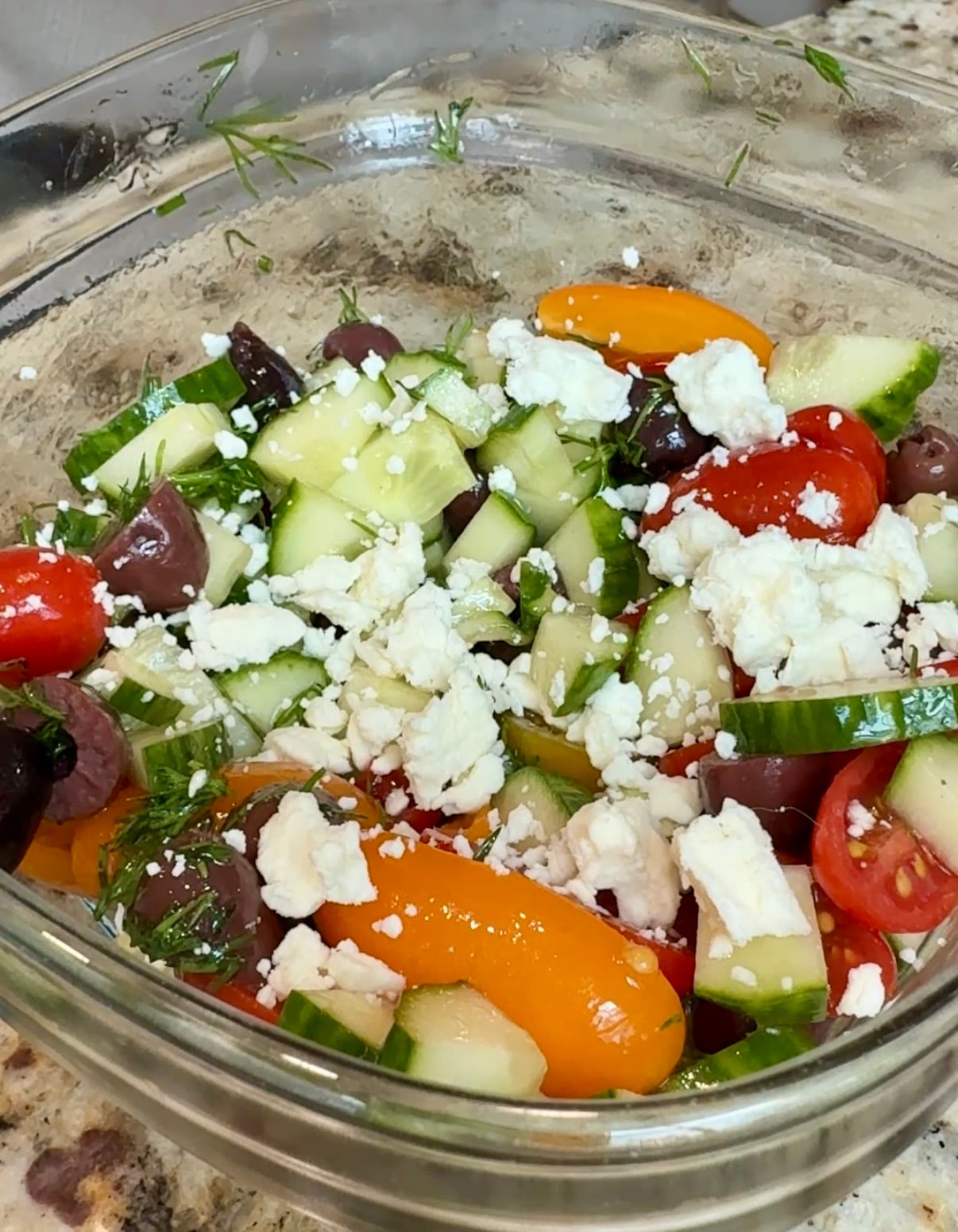 A glass bowl filled with Greek salad featuring chopped cucumbers, cherry tomatoes, kalamata olives, crumbled feta cheese, and fresh herbs on a marble countertop.