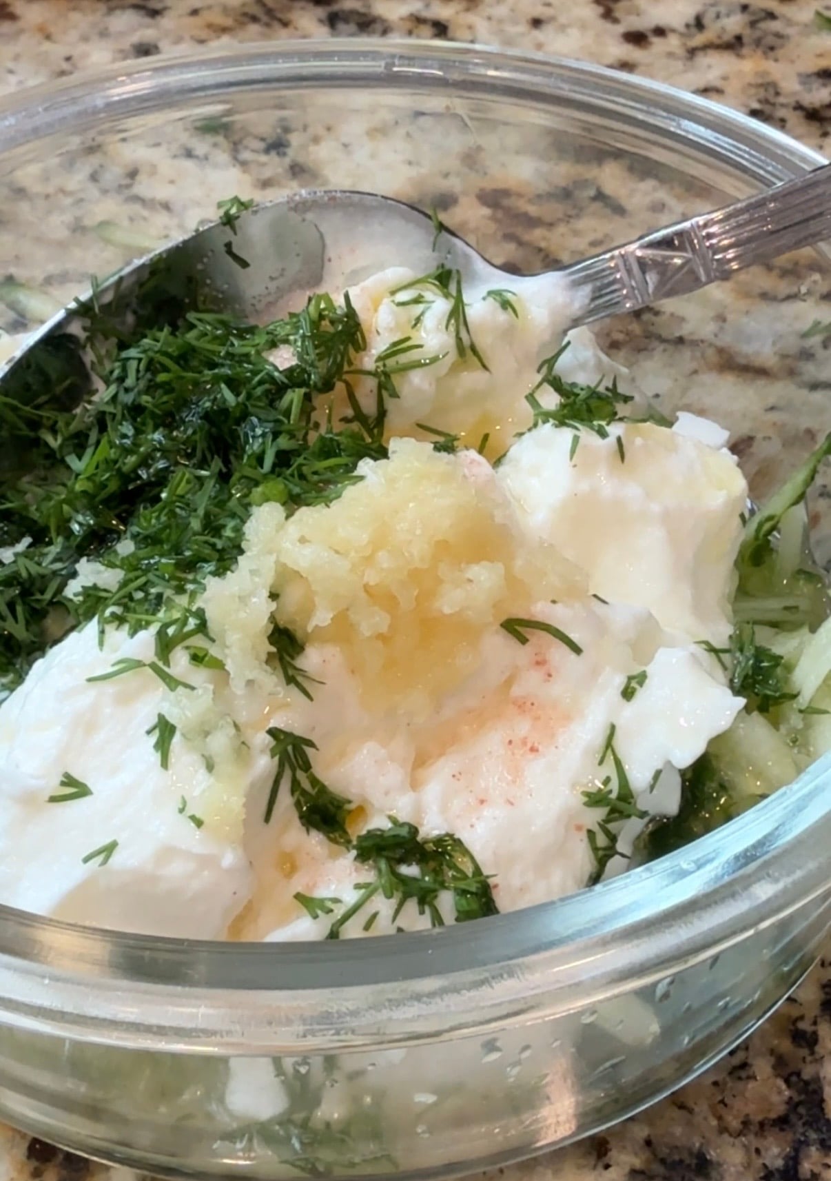 A glass bowl with yogurt, chopped dill, minced garlic, and spices being mixed with a spoon on a granite countertop.