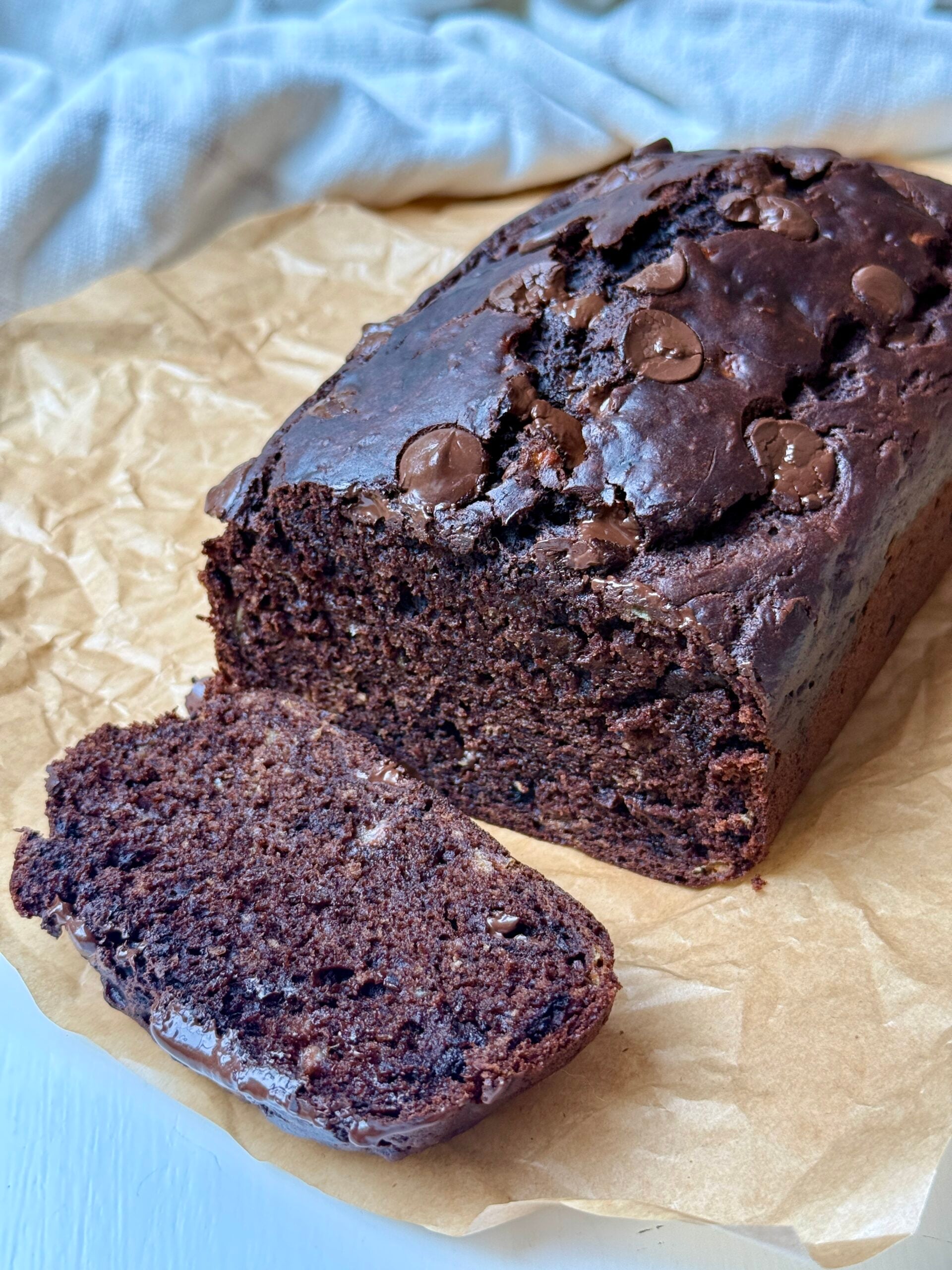 A loaf of chocolate banana bread with chocolate chips sits on brown parchment paper, with one slice cut and placed in front of the loaf.