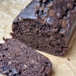 A loaf of chocolate banana bread with chocolate chips sits on brown parchment paper, with one slice cut and placed in front of the loaf.