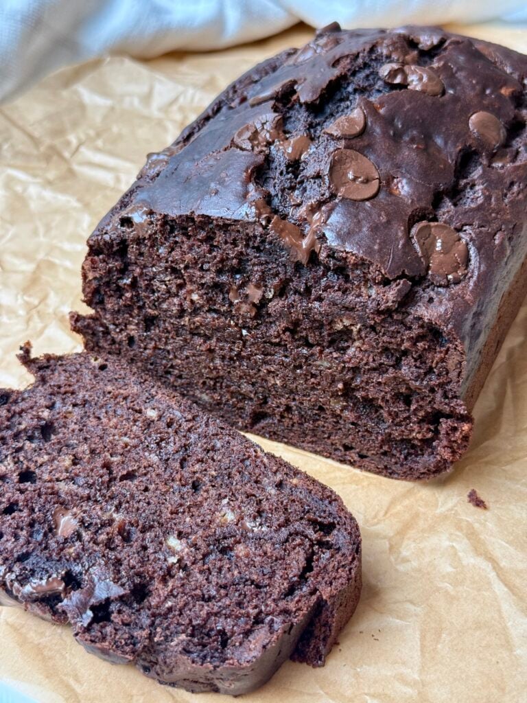 A loaf of chocolate banana bread with chocolate chips sits on brown parchment paper, with one slice cut and placed in front of the loaf.
