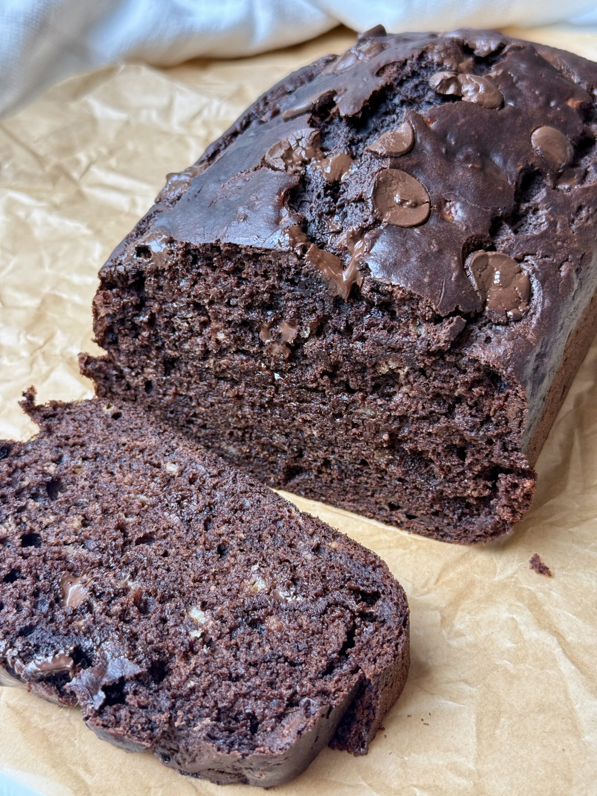 A loaf of chocolate banana bread with chocolate chips sits on brown parchment paper, with one slice cut and placed in front of the loaf.