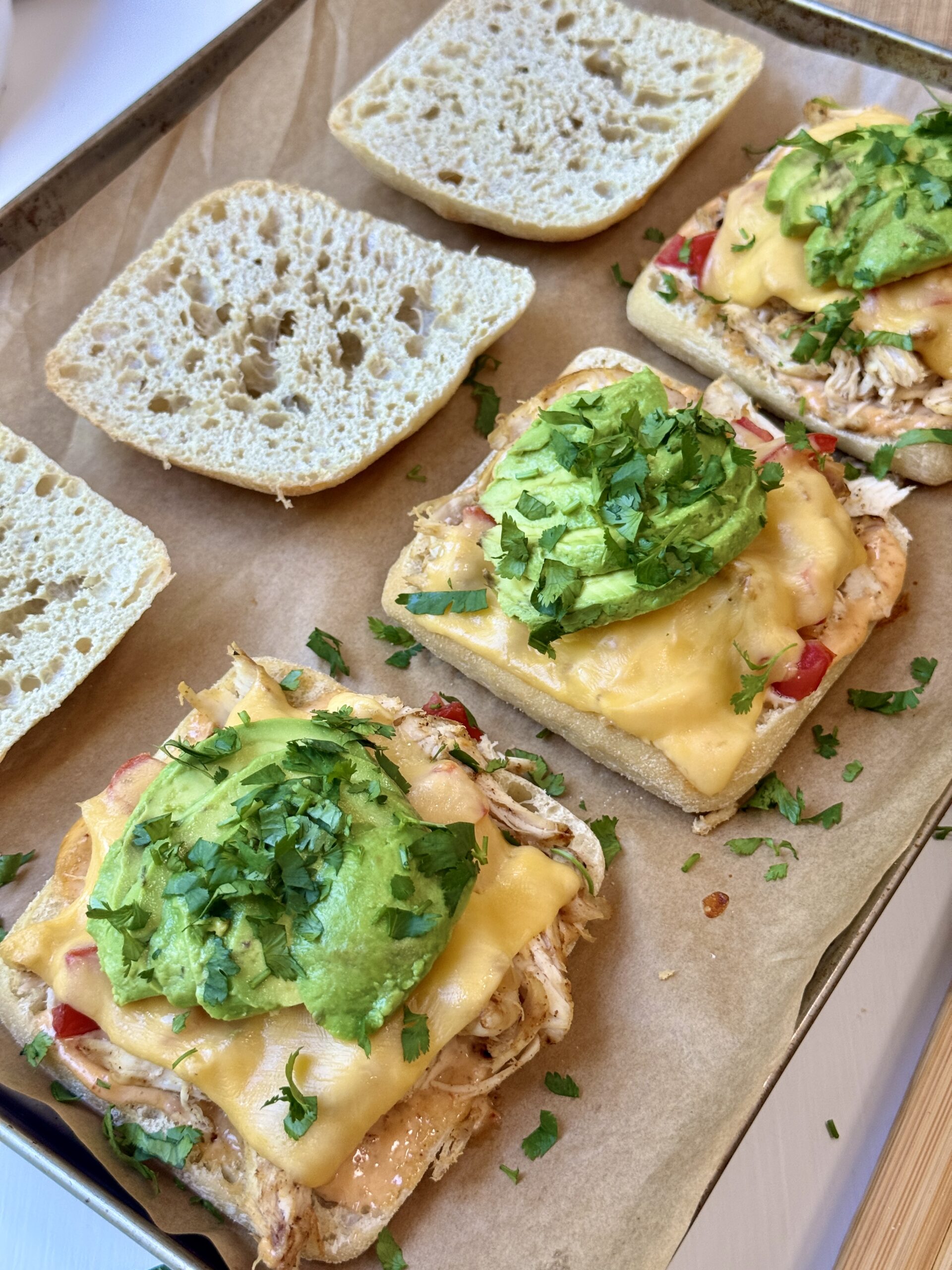 Open ciabatta sandwich buns on a baking sheet. The bottom halves are topped with melted cheese, shredded chicken, sliced avocado, and chopped cilantro. The top halves are plain and set beside them.