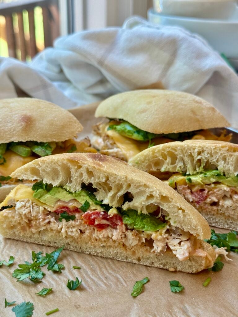 A close-up of ciabatta sandwiches filled with shredded chicken, sliced avocado, melted cheese, and herbs, arranged on parchment paper with chopped cilantro sprinkled around.