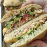 A hand holds a sandwich with shredded chicken, avocado, tomato, herbs, and sauce on ciabatta bread. Other sandwiches are visible on a tray in the background.