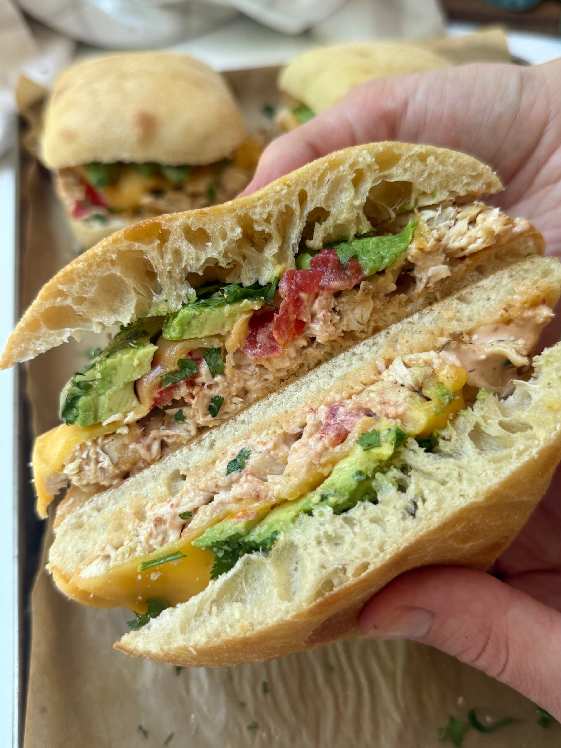 A hand holds a sandwich with shredded chicken, avocado, tomato, herbs, and sauce on ciabatta bread. Other sandwiches are visible on a tray in the background.