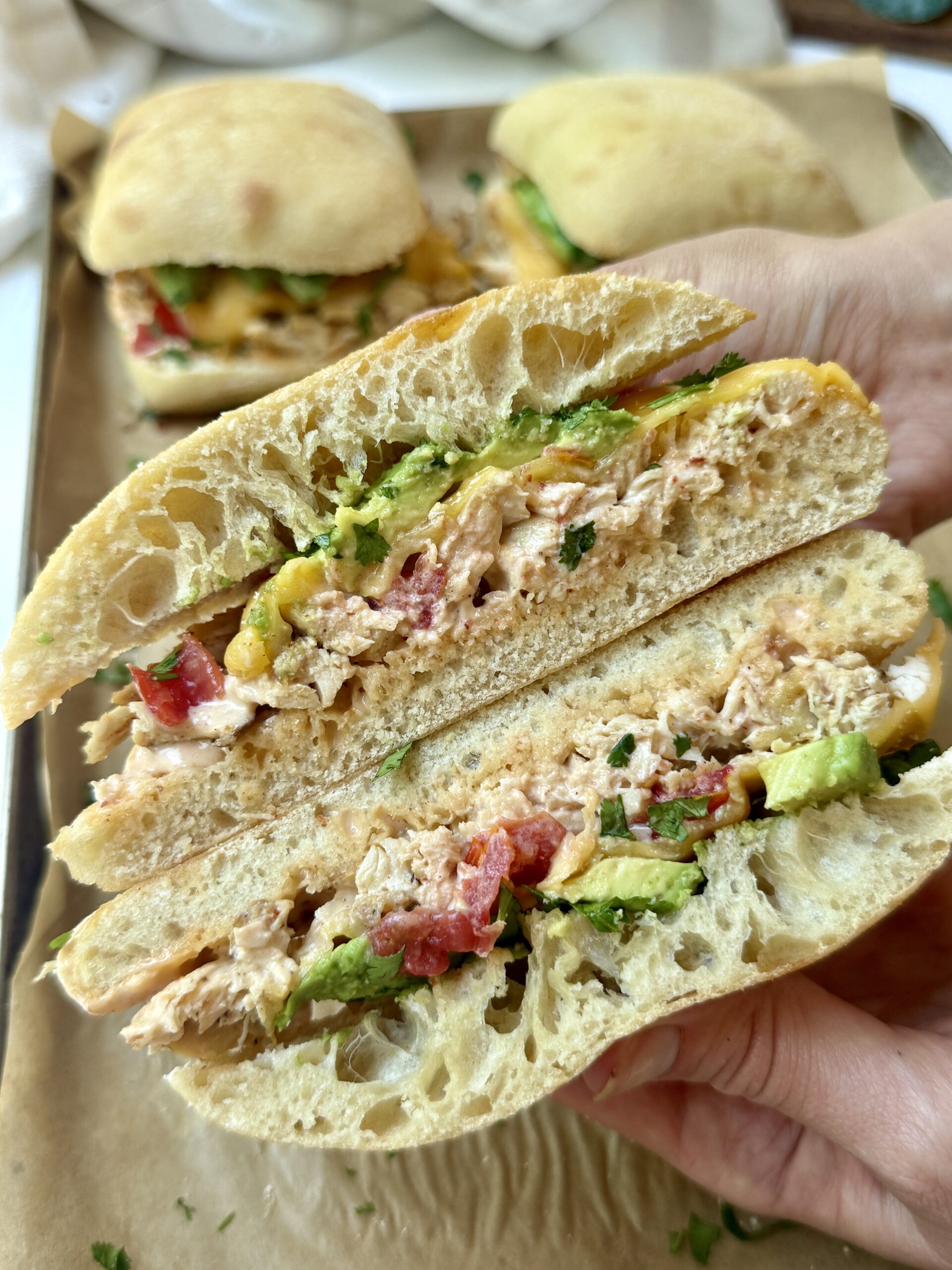 A hand holds a sandwich with shredded chicken, avocado, tomato, herbs, and sauce on ciabatta bread. Other sandwiches are visible on a tray in the background.