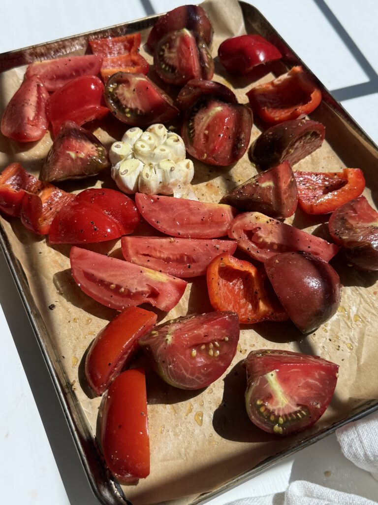 Quartered tomatoes, bell pepper pieces, and a halved garlic bulb on a baking tray lined with parchment paper, glistening in bright sunlight.