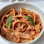 A bowl of ribbon-shaped pasta in creamy tomato sauce, topped with grated cheese and two fresh basil leaves, sits on a white surface with a light cloth in the background.