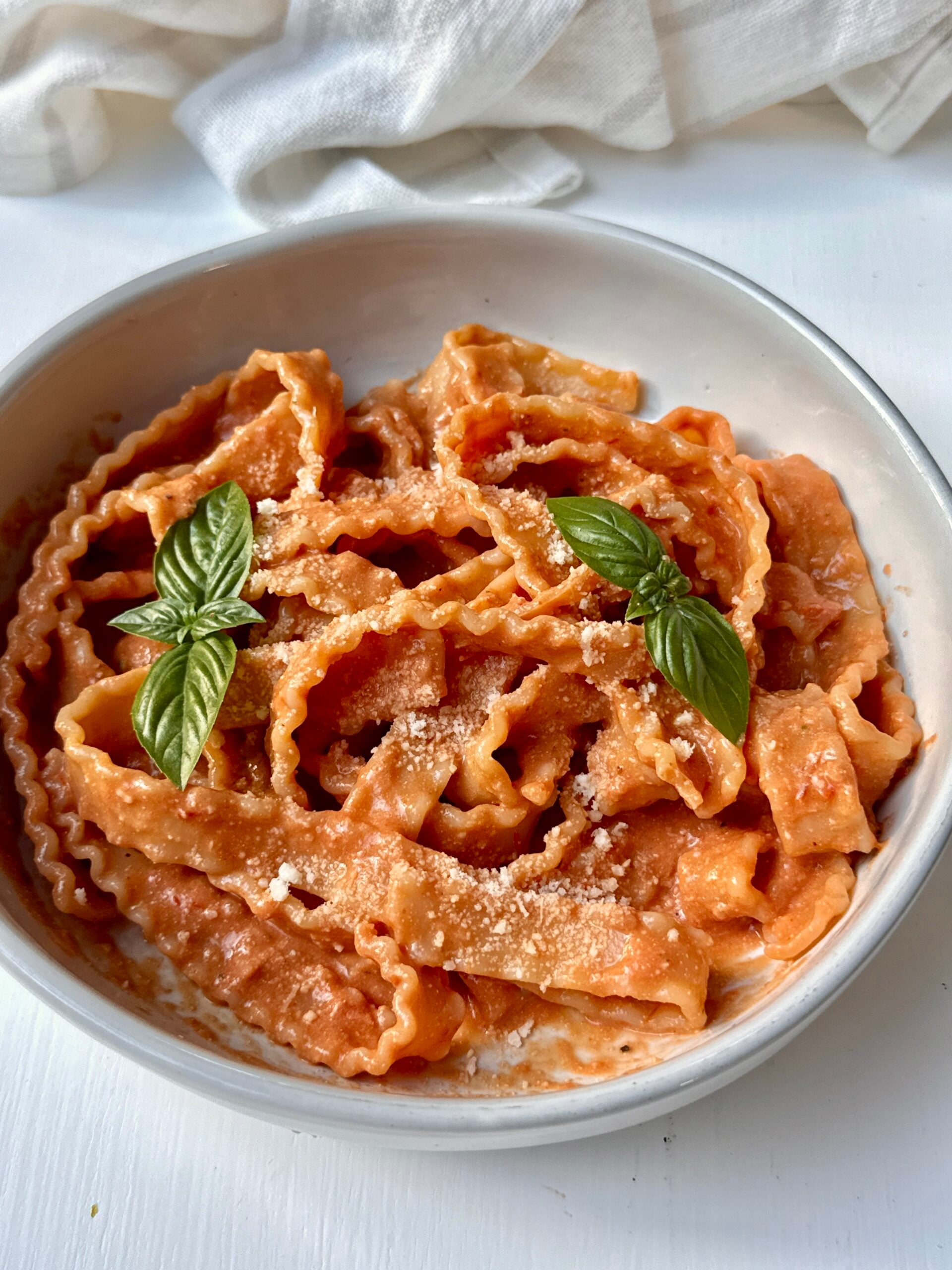 A bowl of ribbon-shaped pasta in creamy tomato sauce, topped with grated cheese and two fresh basil leaves, sits on a white surface with a light cloth in the background.