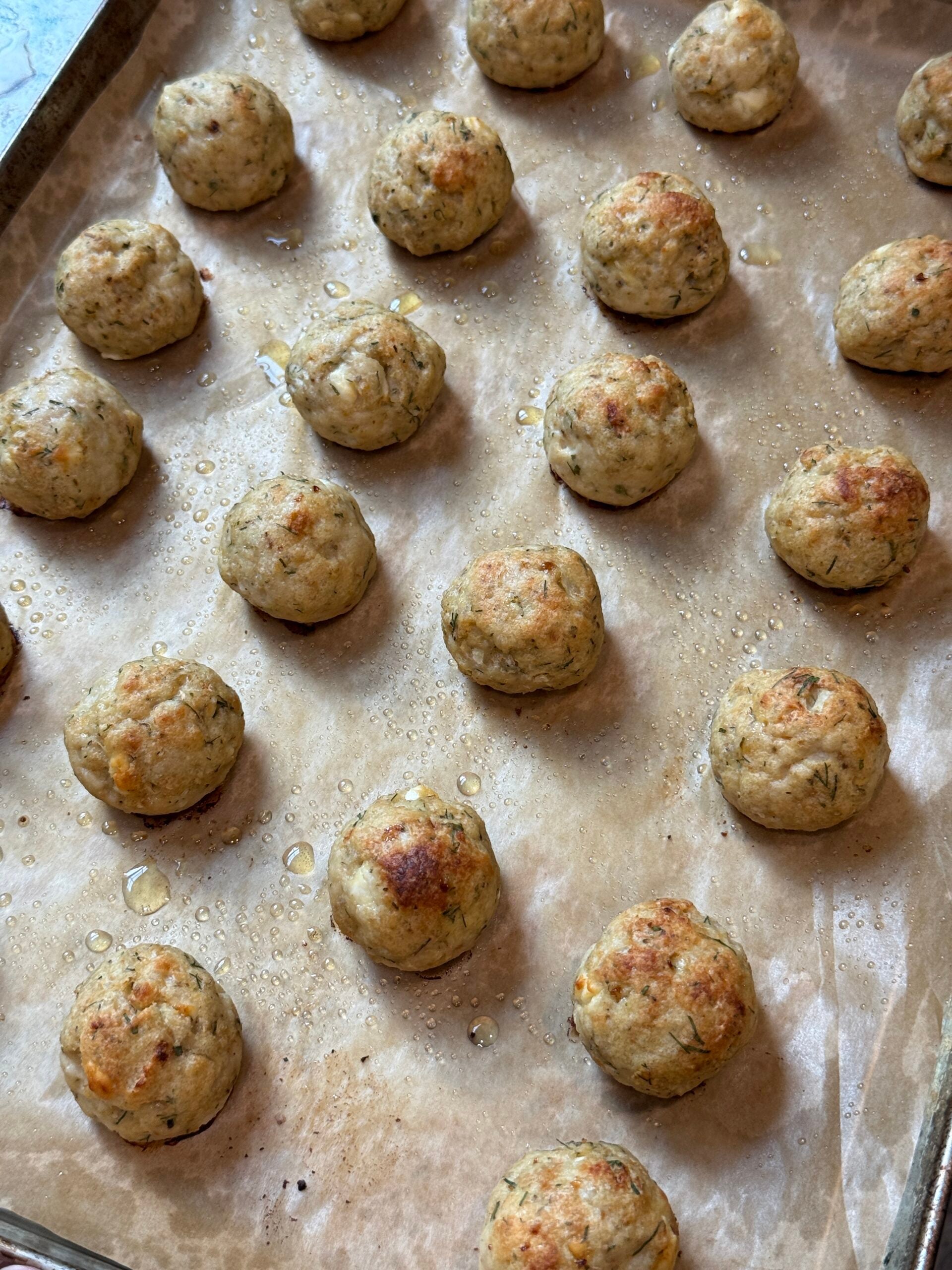 A baking sheet lined with parchment paper holds rows of evenly spaced, golden-brown meatballs, glistening with oil.