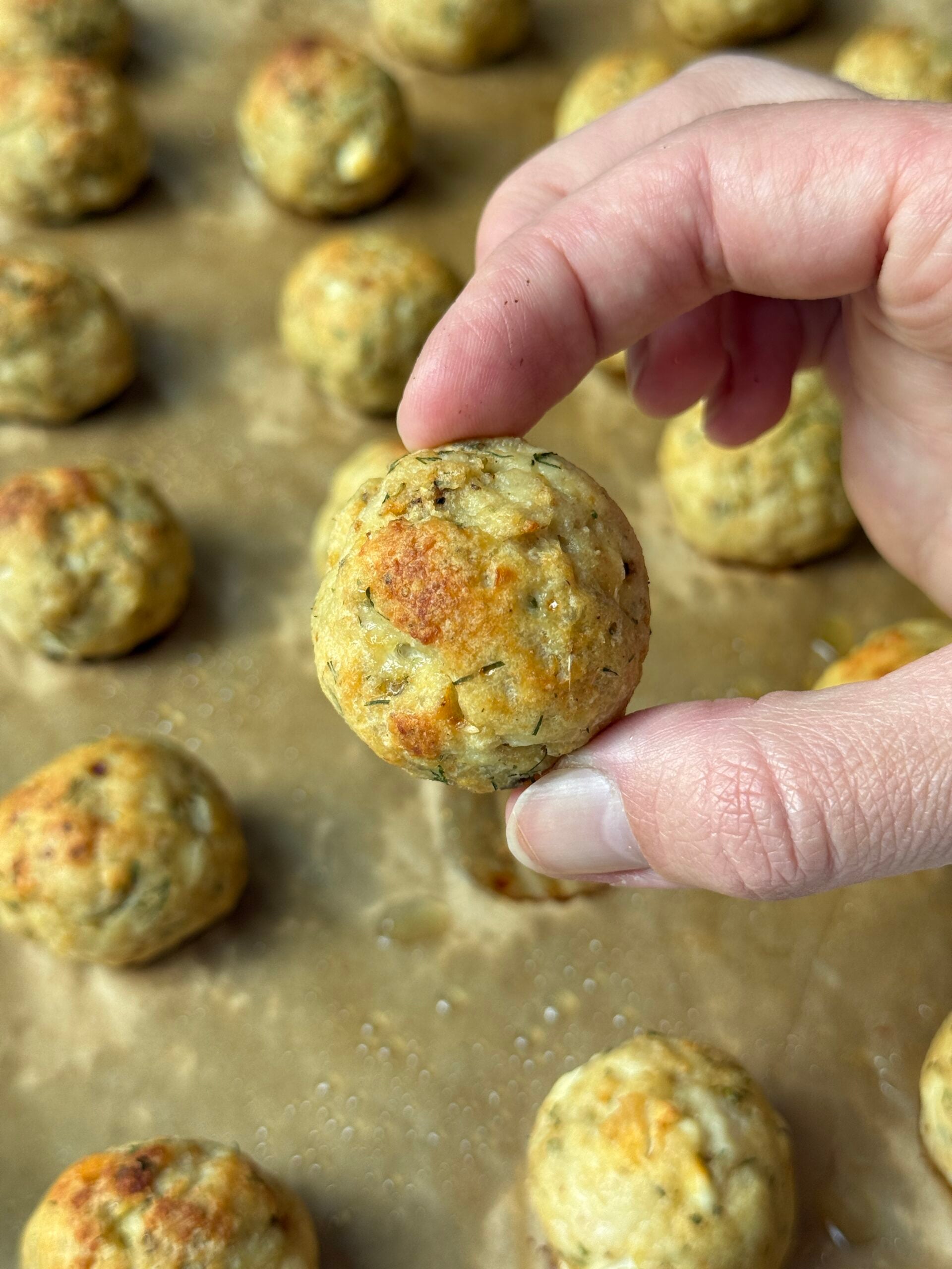 A hand holds a golden-brown baked ball above a parchment-lined tray filled with similar balls, suggesting a homemade savory snack or appetizer.