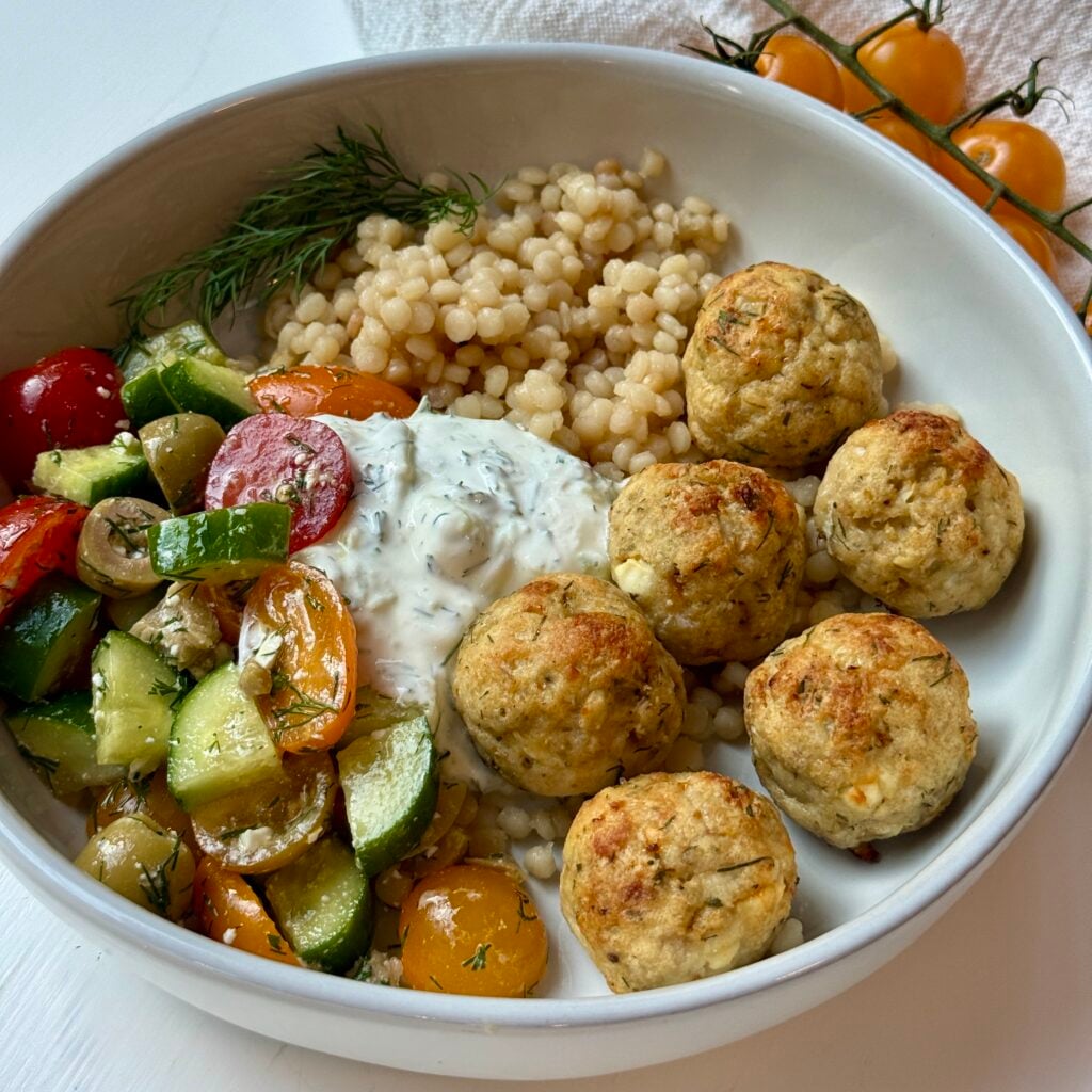 A bowl with couscous, chicken meatballs, tzatziki sauce, and a colorful salad of cherry tomatoes, cucumbers, olives, and herbs. Yellow tomatoes on the vine and fresh dill garnish the dish.