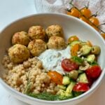 A bowl with couscous, herbed meatballs, cucumber-tomato-olive salad, and a dollop of tzatziki, garnished with dill. Yellow cherry tomatoes are in the background.