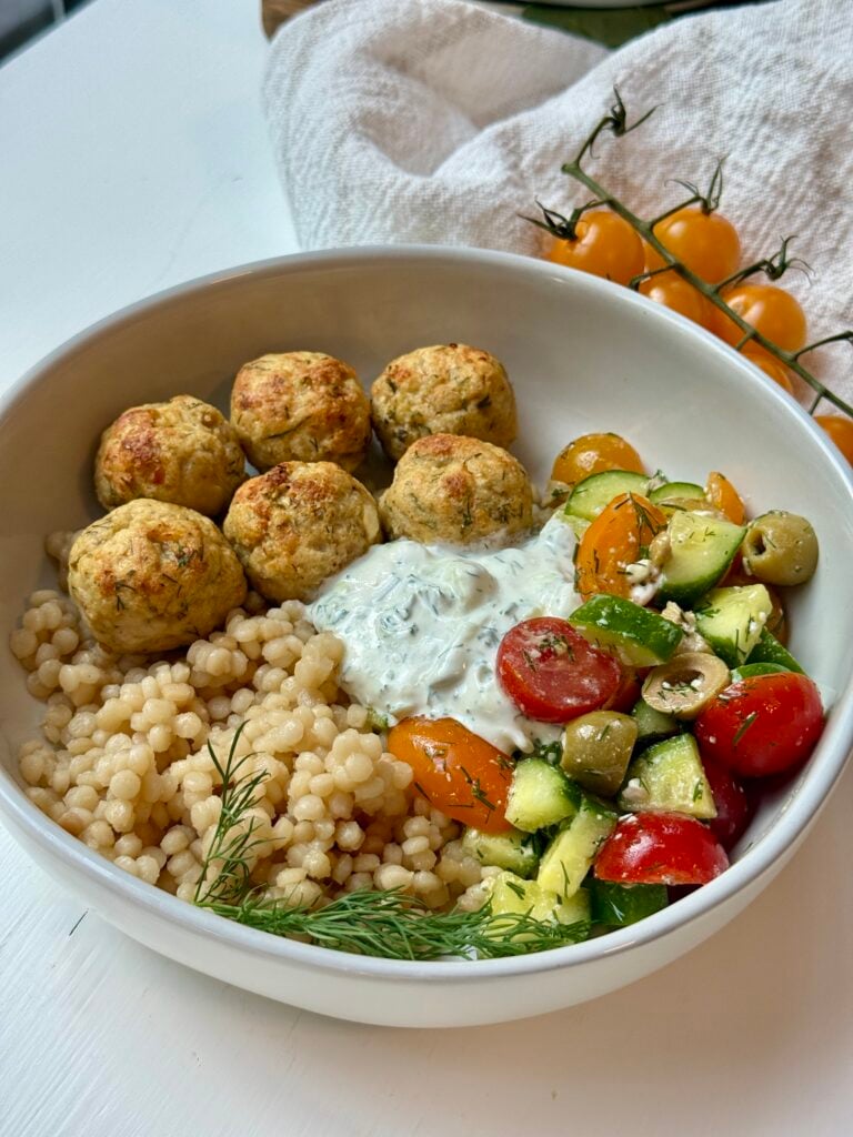 A bowl with couscous, herbed meatballs, cucumber-tomato-olive salad, and a dollop of tzatziki, garnished with dill. Yellow cherry tomatoes are in the background.