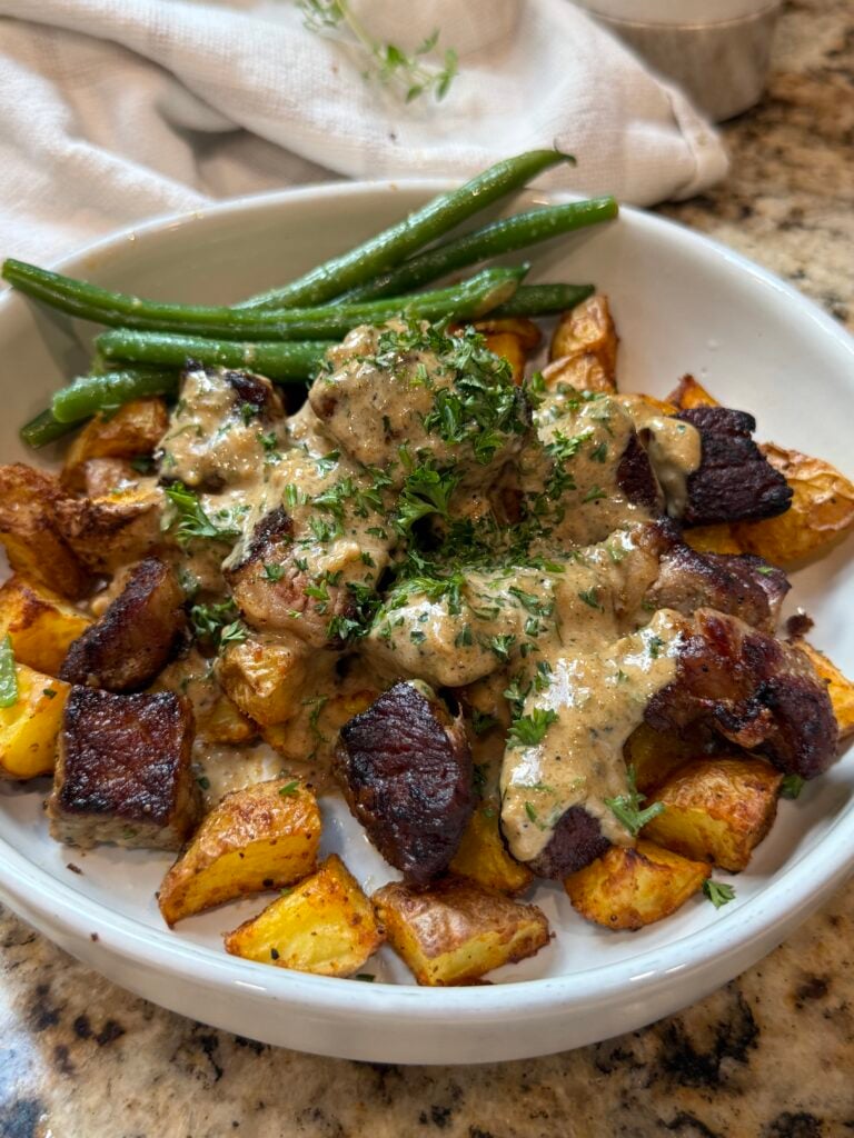 A white bowl filled with roasted potatoes, seared beef pieces topped with creamy sauce and herbs, and green beans on the side.