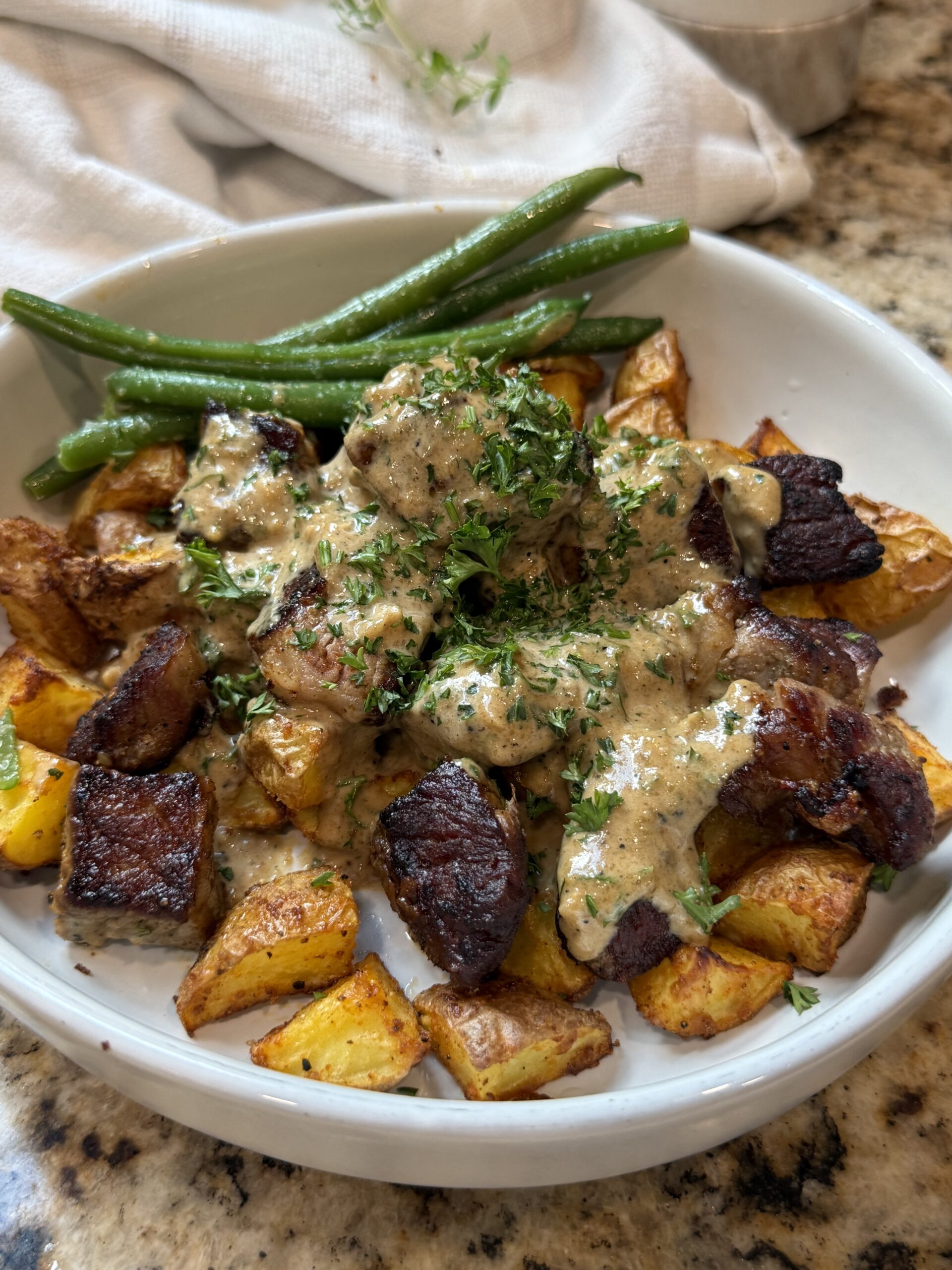 A white bowl filled with roasted potatoes, seared beef pieces topped with creamy sauce and herbs, and green beans on the side.