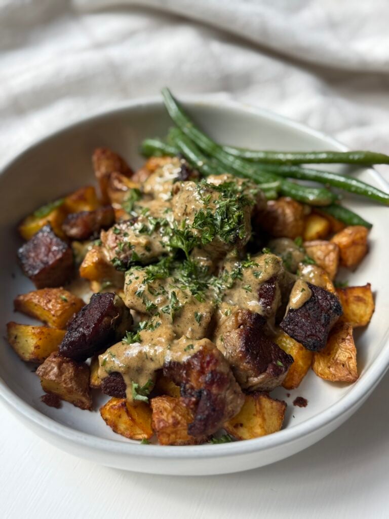 A white bowl filled with roasted potatoes, seared beef pieces topped with creamy sauce and herbs, and green beans on the side.