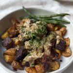 A white bowl filled with roasted potatoes, seared beef pieces topped with creamy sauce and herbs, and green beans on the side.