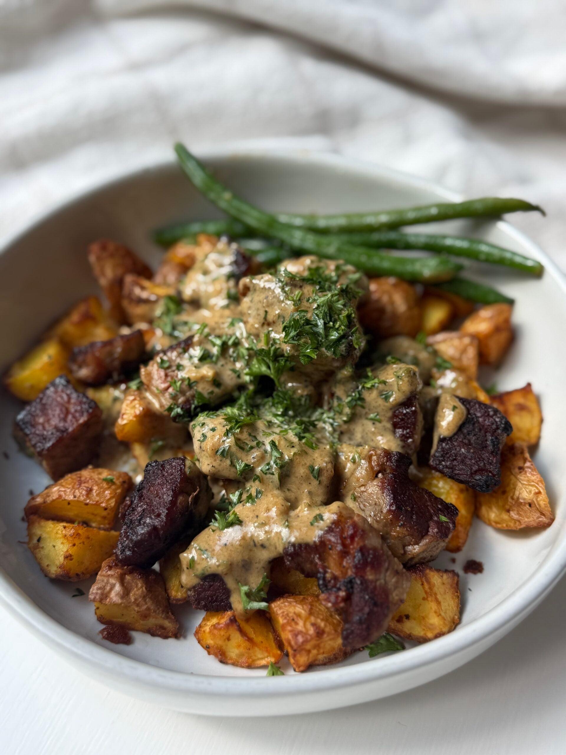 A white bowl filled with roasted potatoes, seared beef pieces topped with creamy sauce and herbs, and green beans on the side.