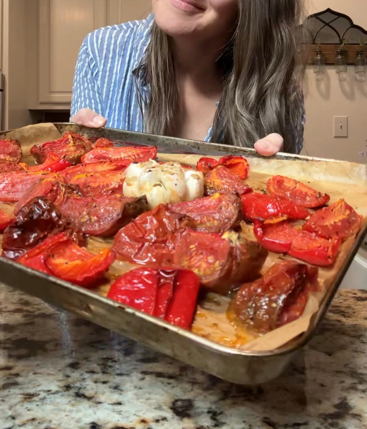 A woman in a striped shirt holds a tray of roasted tomatoes, peppers, and garlic on parchment paper in a kitchen.