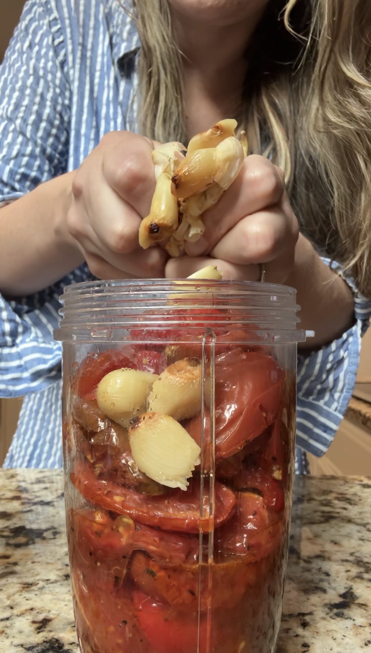 A person in a blue-striped shirt squeezes roasted garlic cloves into a blender cup filled with roasted tomatoes and other vegetables on a kitchen counter.