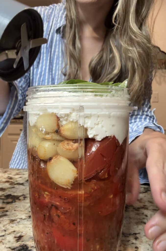 A woman prepares to blend roasted tomatoes, garlic, feta cheese, olive oil, and fresh basil in a blender cup on a kitchen counter.