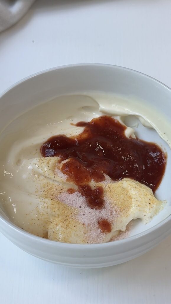 A white bowl containing creamy white yogurt, a dollop of reddish-brown jam or sauce, and a sprinkling of light brown and pinkish seasonings on top.