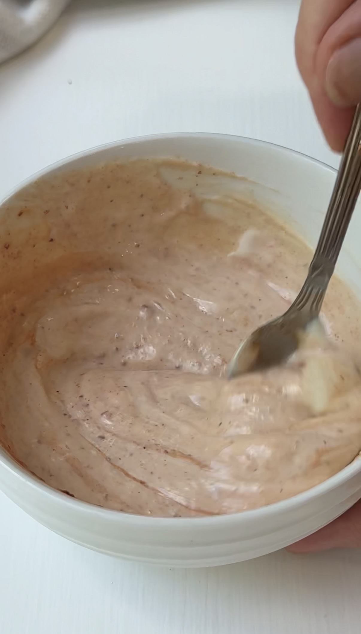 A hand stirs a creamy, light orange sauce with visible spices in a white bowl using a fork.