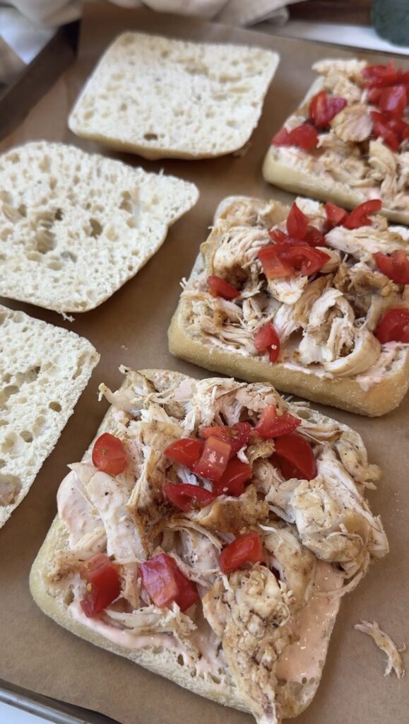 Three open ciabatta rolls topped with shredded chicken, diced tomatoes, and a spread. The top halves of the rolls are placed nearby on parchment paper, ready to be assembled into sandwiches.