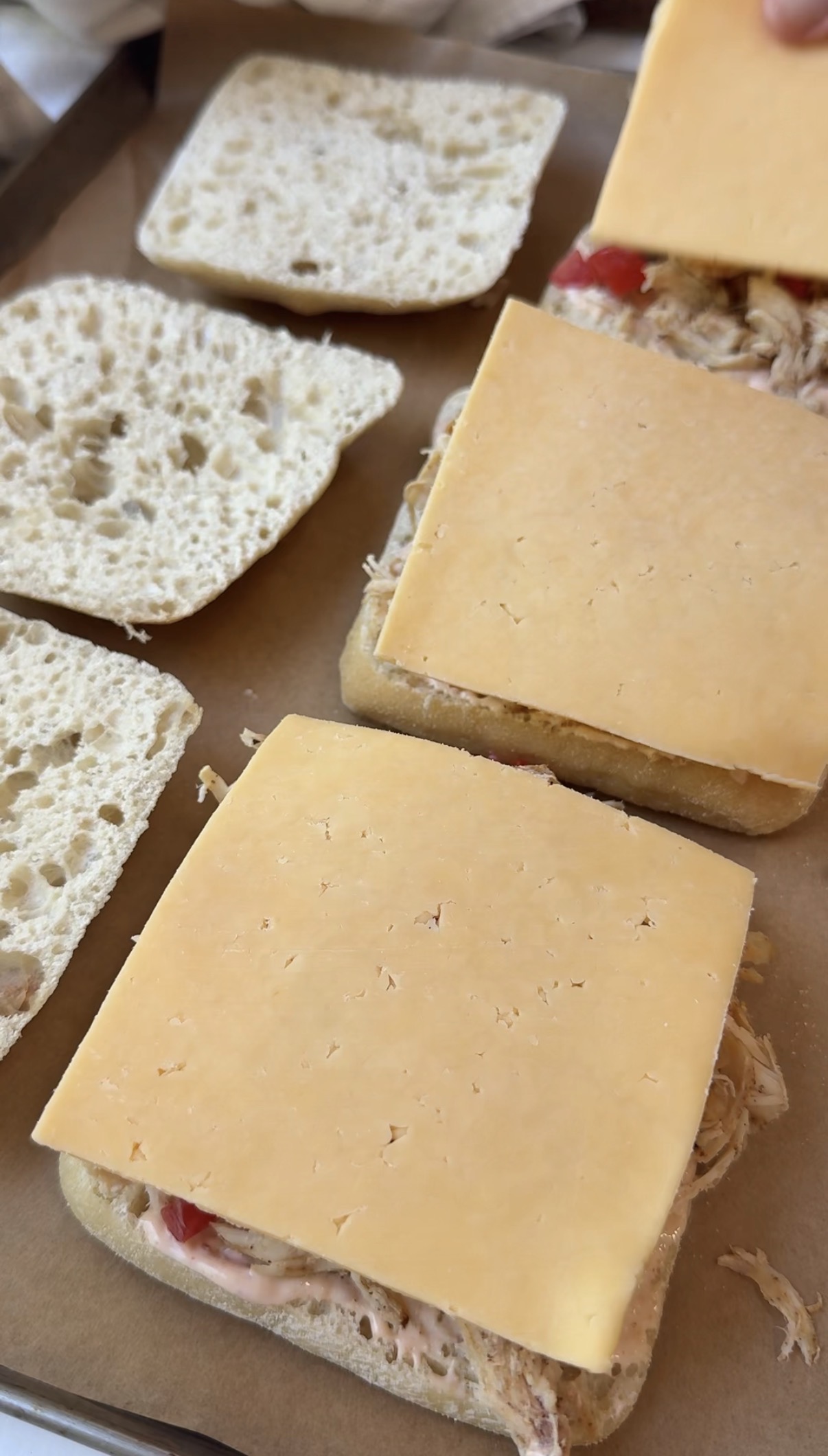Two sandwiches being prepared on a brown surface, with shredded meat, two slices of cheddar cheese, and split ciabatta bread visible.