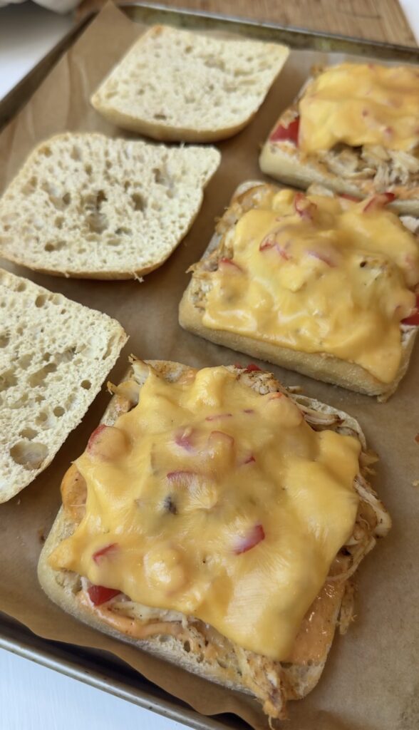 Three open-faced sandwiches with melted cheese and shredded chicken on square buns sit on parchment paper, with top halves of the buns placed nearby.