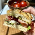 A hand holds a sandwich with focaccia bread, prosciutto, mozzarella, lettuce, and pesto. In the background, tomatoes sit in a wooden bowl.