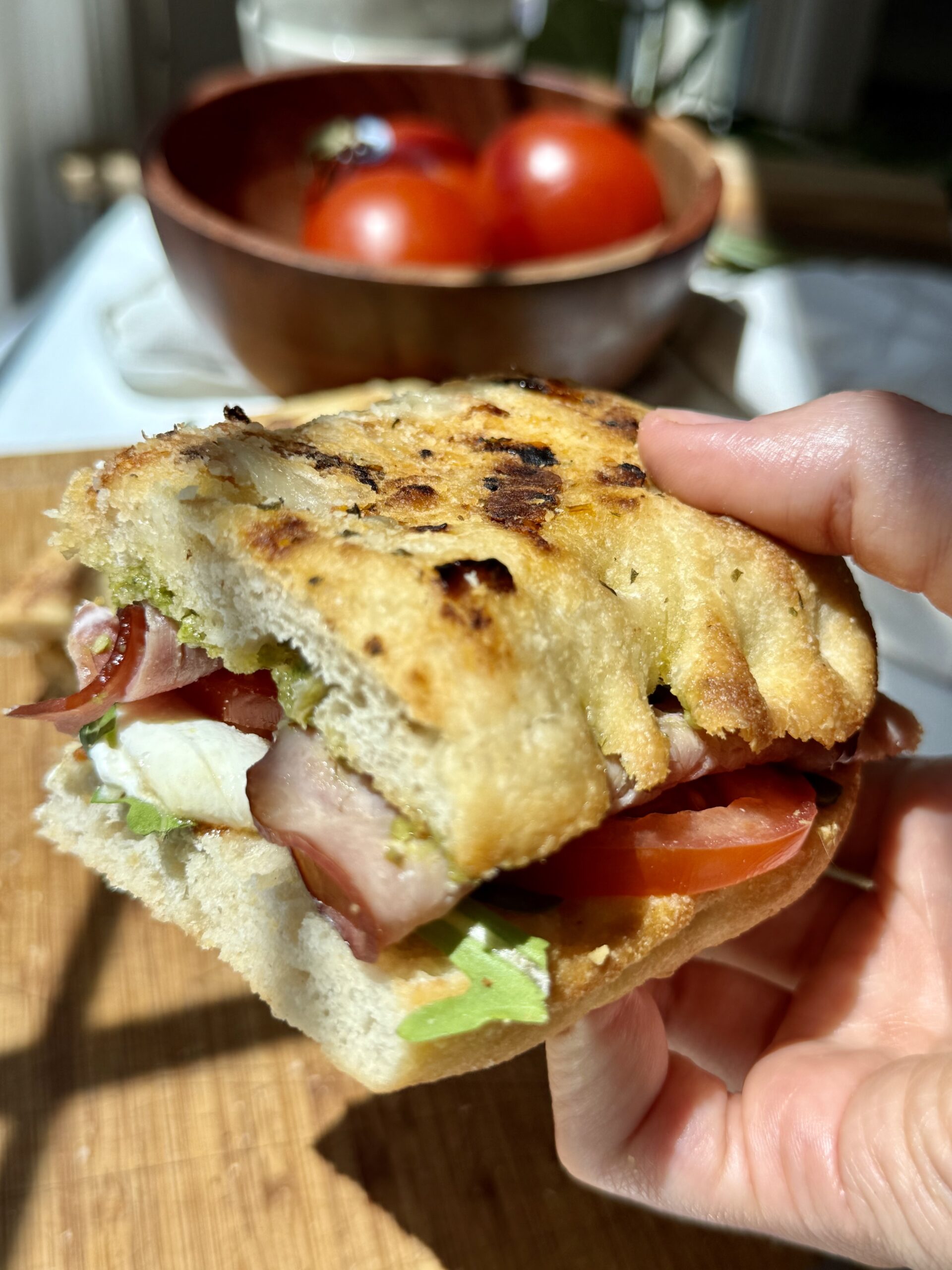 A hand holds a partially eaten sandwich with prosciutto, mozzarella, tomato, and greens on toasted bread. A wooden bowl with tomatoes is in the blurred background.