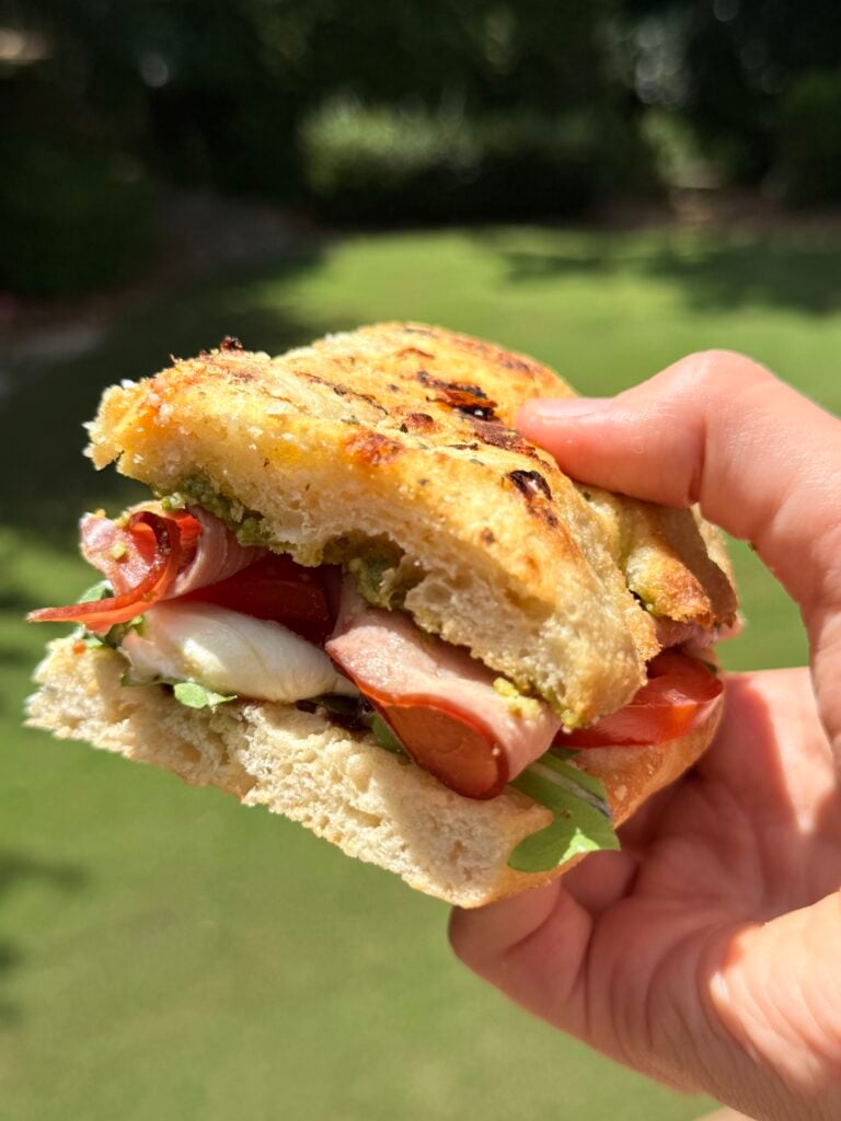 A hand holds a sandwich with toasted bread, lettuce, tomato, cheese, and slices of cured meat. The background is a sunny, grassy outdoor area.